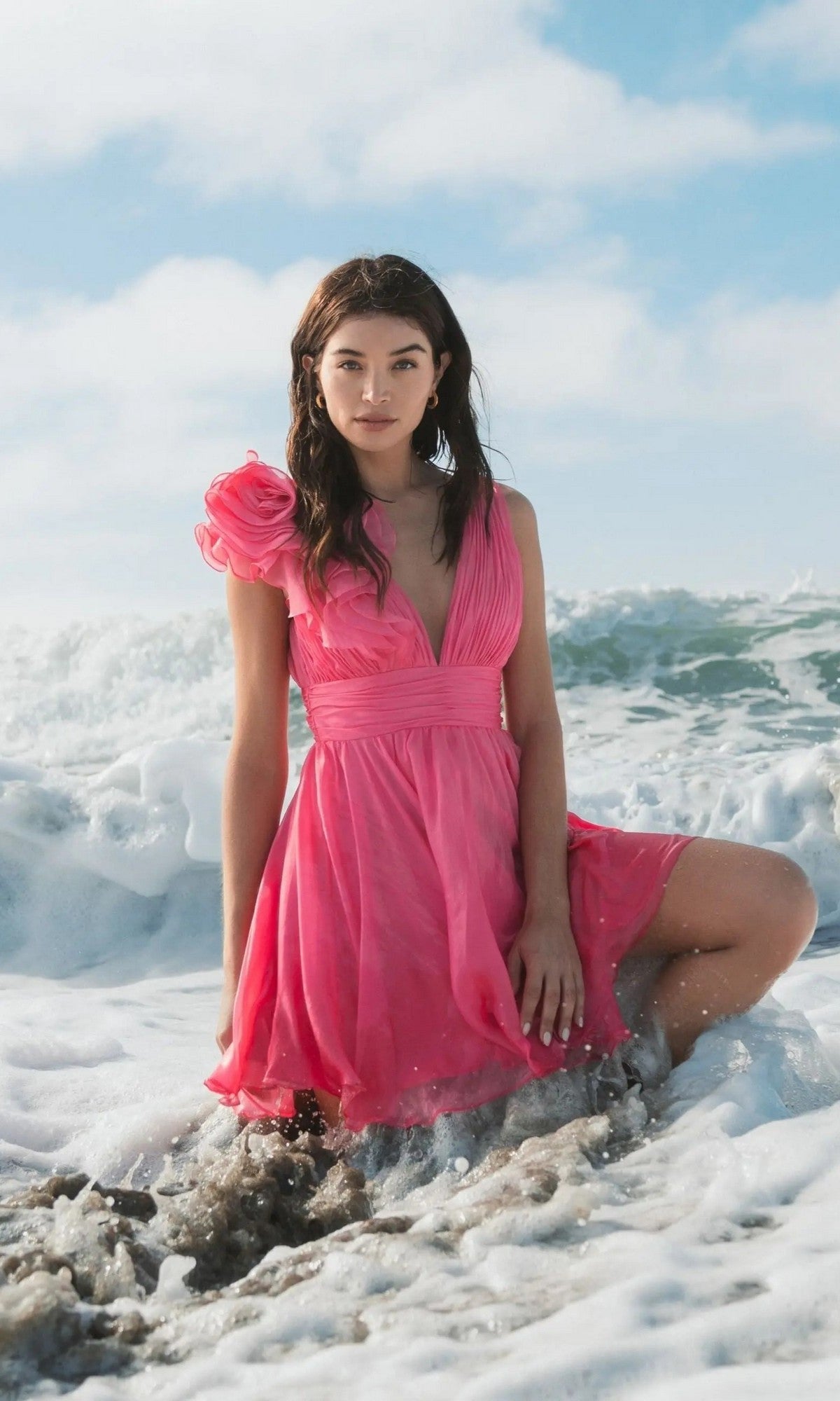 A woman in a Mac Duggal 48952 ruffle short white party dress kneels on wet sand at the beach, with waves and sky in the background.